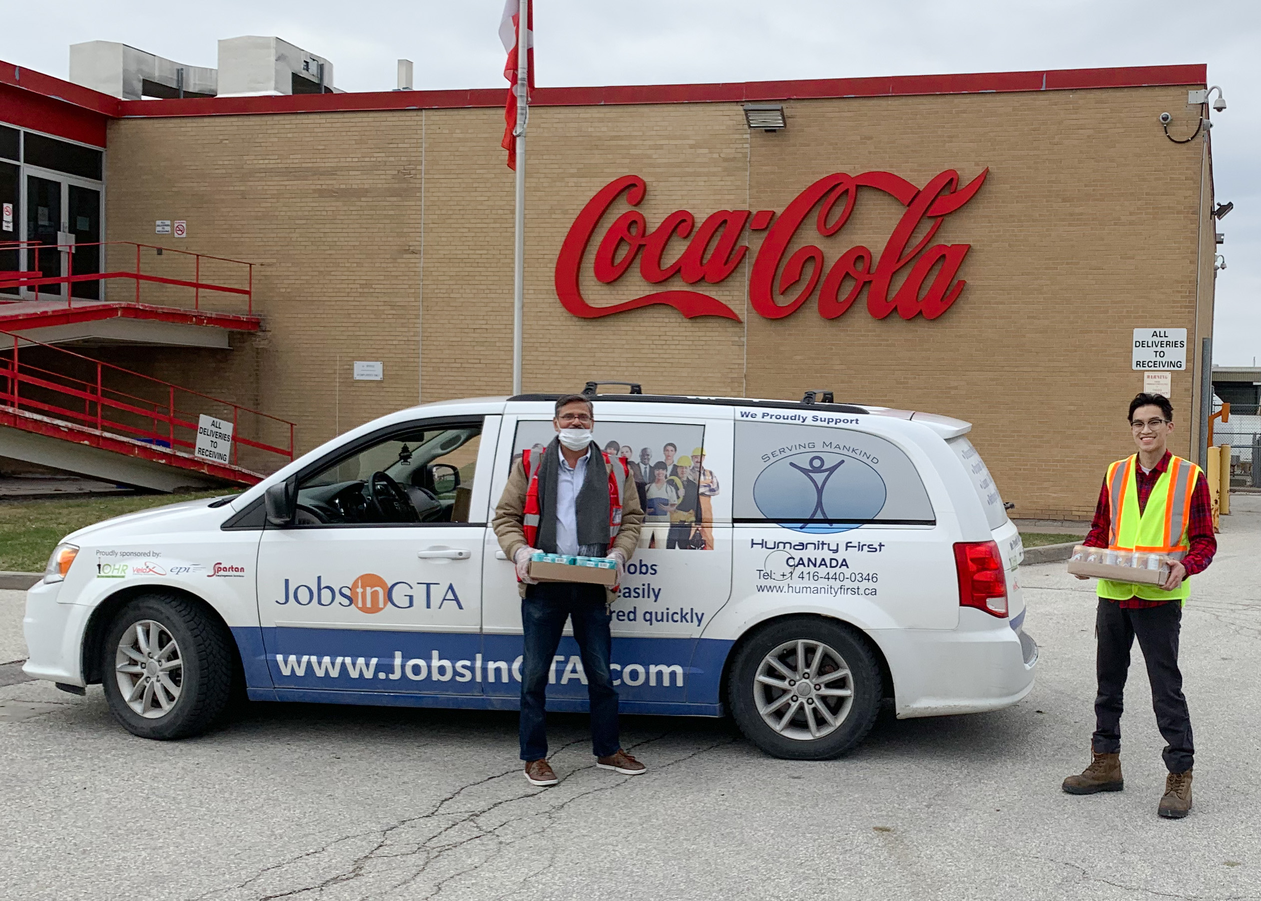 Deux hommes debout devant une camionnette Jobs in GTA, souriant à la caméra et tenant chacun une boîte de produits Coca-Cola. Un bâtiment en briques avec un grand logo Coca-Cola rouge se dresse en arrière-plan