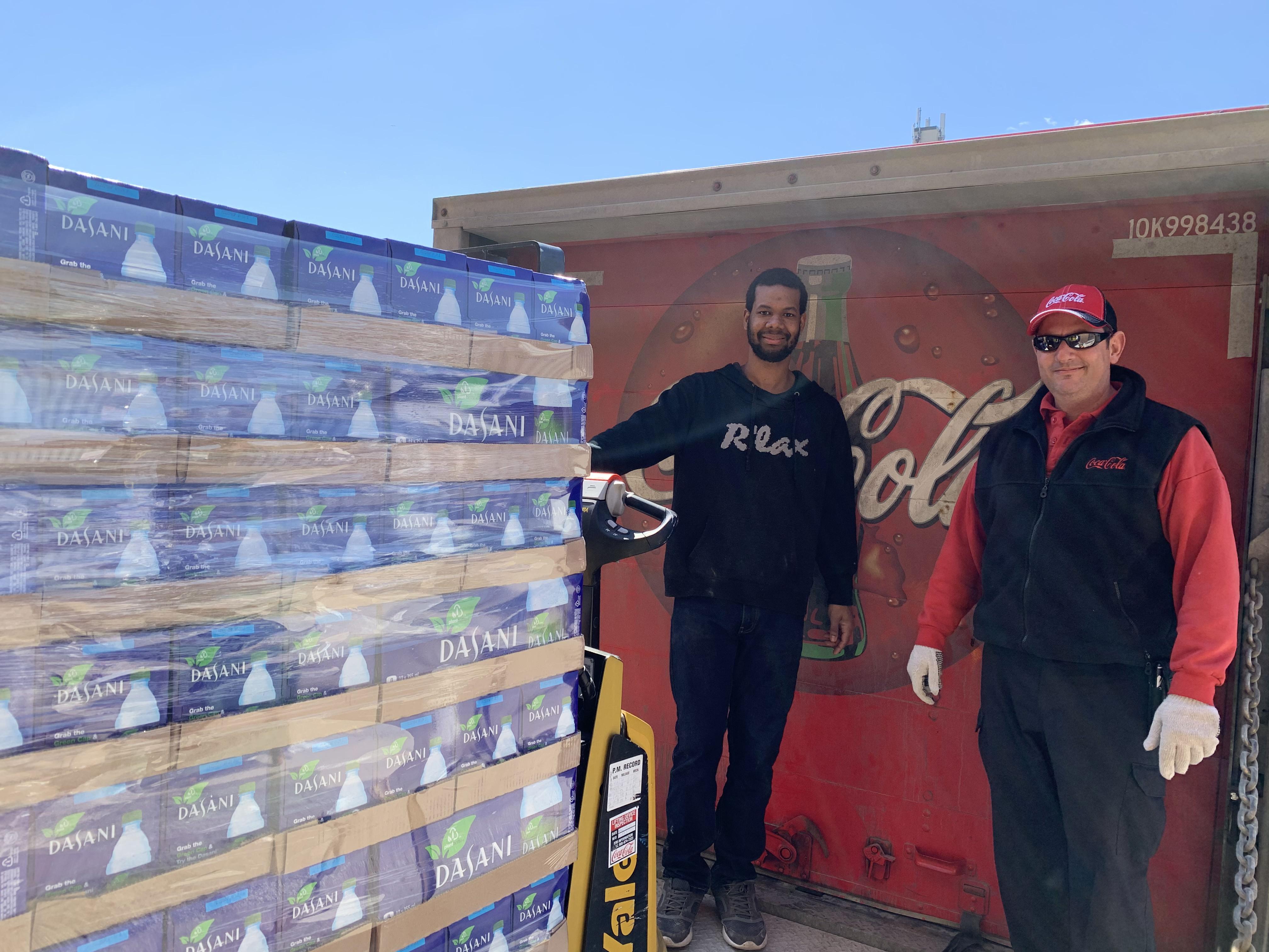 Deux hommes se tiennent sur un quai de chargement devant un camion Coca-Cola, un transpalette avec une pile de palettes d'eau Dasani est au premier plan